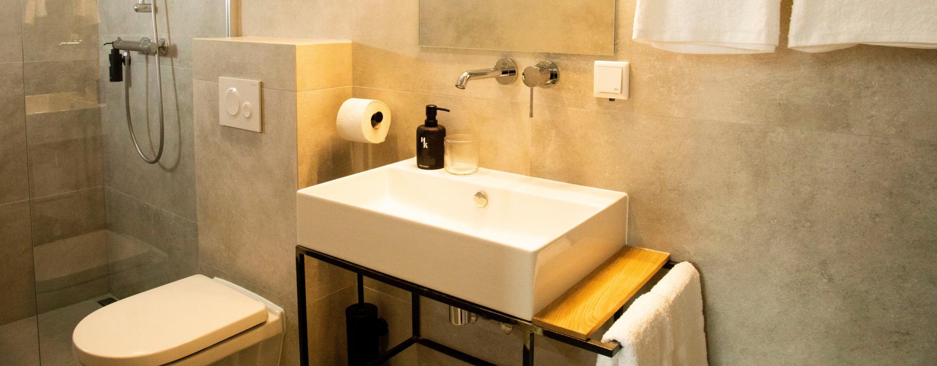 Een moderne badkamer met grote grijze tegels, een ruime douche, een vierkante wasbak, een toilet en witte handdoeken